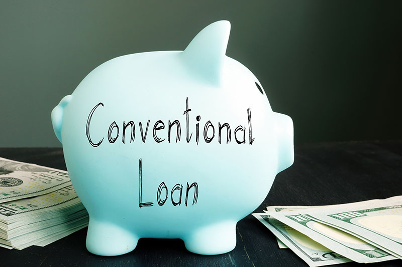 A light blue piggy bank labeled Conventional Loan sits on a table next to stacks of U.S. dollar bills, against a dark background.