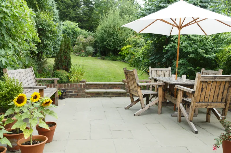 A patio with wooden benches and a dining table set beneath a large white umbrella, surrounded by potted sunflowers and lush green garden foliage in the background.